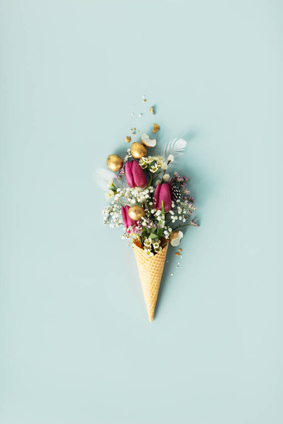 Velikonoční kompozice. Zmrzlinový kužel s krásnými květy a zlatými velikonočními vejci na modrém pozadí ploché snášky - Fotografie, Obrázek