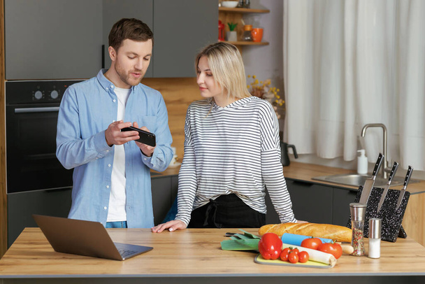 Усміхнена любляча пара готує салат разом, стоячи на кухні вдома і використовуючи мобільний телефон і ноутбук
 - Фото, зображення