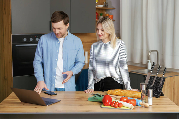 Agradable pareja familiar de pie cerca de una gran mesa de madera en la cocina moderna, mirando a la pantalla del ordenador portátil. Feliz joven pareja empezando a preparar el almuerzo - Foto, imagen