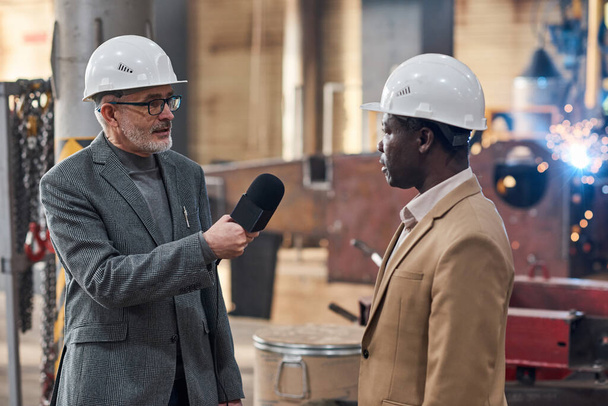 Sert başlıklı Afrikalı işadamı mikrofonla gazeteciye röportaj veriyor ve haber sırasında fabrikadaki durum hakkında konuşuyor - Fotoğraf, Görsel