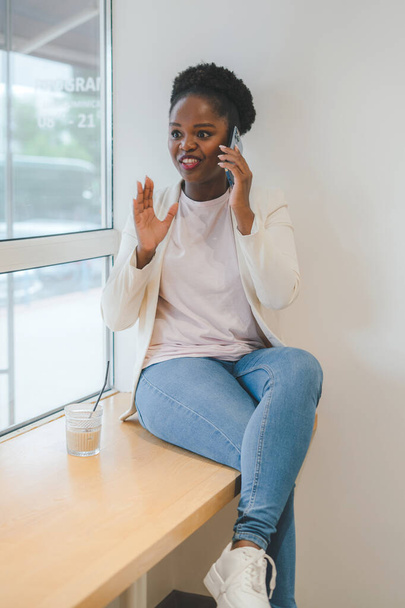Kısa siyah kıvırcık saçlı Afro-Amerikalı bir kadın bir kafede cep telefonuyla oturuyor. Kavram: telefonu kullanmak. Cep telefonu iletişimi. - Fotoğraf, Görsel