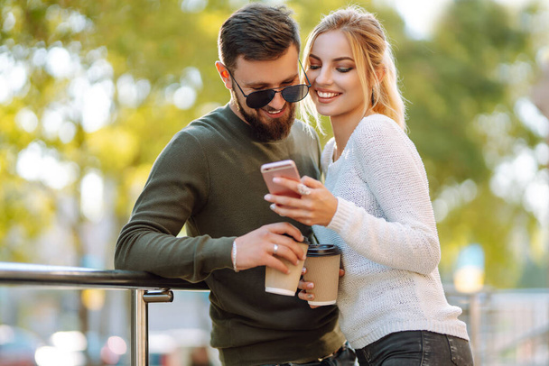 Young couple having fun talking, drinking coffee and using the phone while walking in the morning city. Relaxation, youth, love, lifestyle, selfie, blogging. - 写真・画像