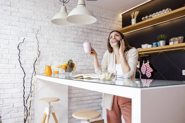 Mujer sonriente comiendo alimentos saludables mientras usa su teléfono móvil en casa. Chica hablando por teléfono en la cocina. - Foto, imagen