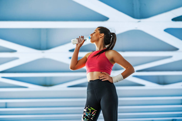 Attractive girl in sportswear drinking water after workout on city bridge. - Photo, Image