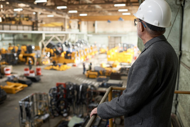 Вид спереду зрілого інженера в робочому шоломі стоячи на балконі і спостерігаючи за виробництвом на заводі
 - Фото, зображення