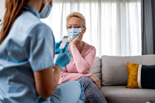 Seniorin trägt Mundschutz, während Arzt bei Hausbesuch Temperatur misst. - Foto, Bild