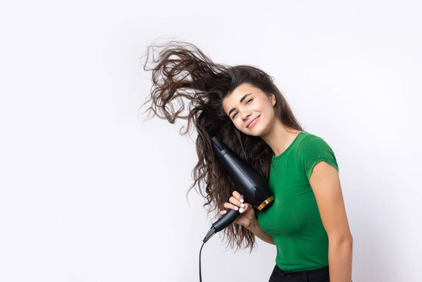 A cute young girl dressed in a green top dries her beautiful long silky hair with a hair dryer against a white background. - Foto, Imagen