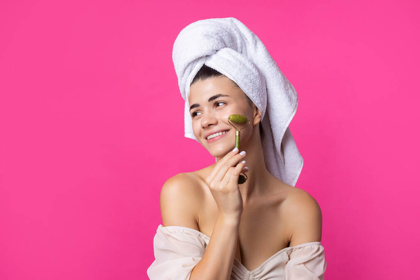 A beautiful cheerful attractive girl with a towel on her head holds a cosmetic roller near her face against a pink background. - Fotoğraf, Görsel