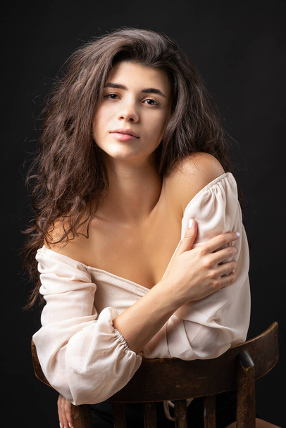 Classic studio portrait of a young brunette in a white loose top, who is sitting on a chair against a black background. - Photo, Image