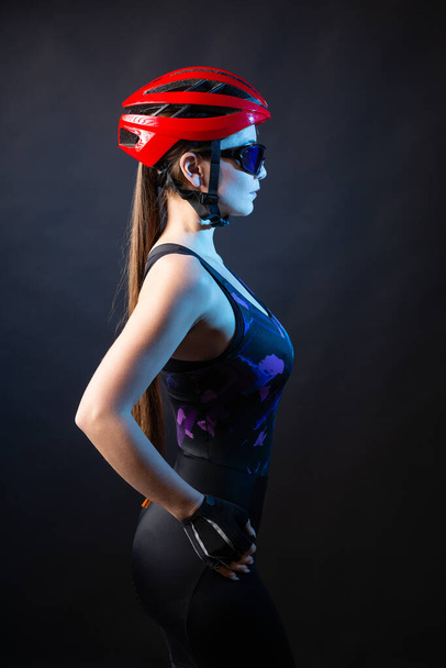 A young female cyclist wearing a safety helmet and glasses, dressed in a bib shorts poses against a black background in the studio. - Foto, afbeelding