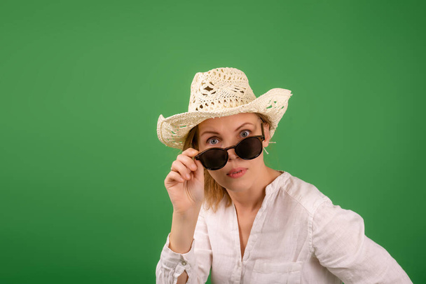 Cheerful amazed woman in a hat and sunglasses in a white shirt on a green background. Looks into the camera with his glasses down. - Foto, afbeelding