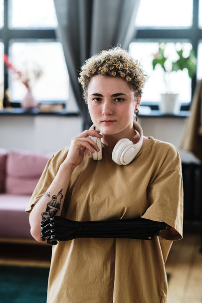 Retrato de mujer joven con brazo protésico escuchando música en auriculares inalámbricos de pie en la habitación - Foto, imagen