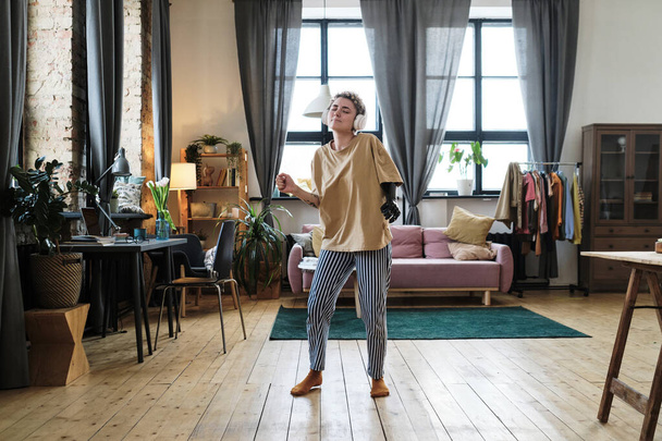 Mujer joven con brazo protésico bailando mientras escucha música en auriculares inalámbricos en la sala de estar en casa - Foto, imagen