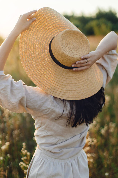 Femme boho élégant avec chapeau de paille posant parmi les fleurs sauvages dans la lumière du coucher du soleil, vue de dos. Délice d'été et tranquillité. Jeune femelle insouciante en lin rustique relaxant dans la prairie d'été - Photo, image