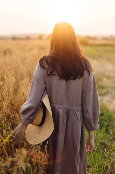 Stílusos nő szalmakalapban sétál a zab mezőn napnyugtakor, visszapillantással. Légköri nyugalmi pillanat. Fiatal nő vászon ruha pihentető este nyári táj, vidéki lassú élet - Fotó, kép