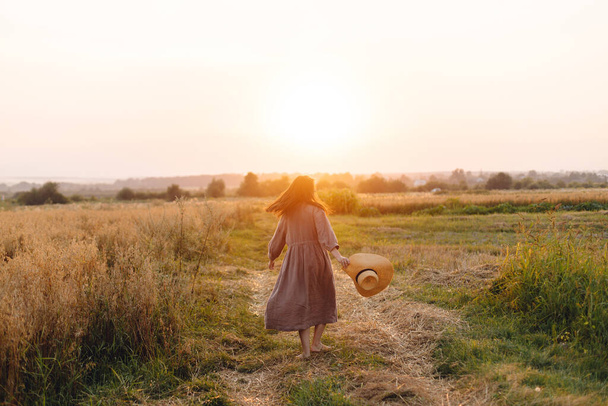 Stylowa kobieta w słomkowym kapeluszu tańcząca na polu owsianym w świetle zachodu słońca. Atmosferyczny szczęśliwy moment. Młoda kobieta w rustykalnej sukni lnianej relaks w lecie wsi, życie na wsi - Zdjęcie, obraz