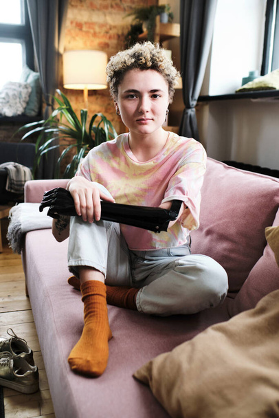 Πορτρέτο του κοριτσιού με προσθετικό χέρι σε casual ρούχα κάθεται στον καναπέ στο σαλόνι και κοιτάζοντας κάμερα - Φωτογραφία, εικόνα