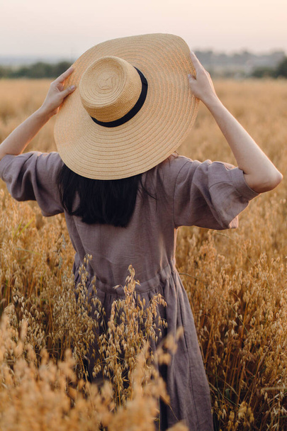 Mujer con estilo con sombrero de paja de pie en el campo de avena en la luz de la noche, vista trasera. Momento de tranquilidad atmosférica. Mujer joven en vestido de lino rústico relajante en el campo de verano. Vida rural lenta - Foto, Imagen