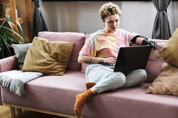 Kolu protez olan genç bir kadın kanepeye oturmuş dizüstü bilgisayarı bacaklarında ve oturma odasında internetten çalışıyor. - Fotoğraf, Görsel