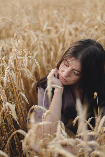 Стильная спокойная женщина, сидящая на пшеничном поле в вечернем свете. Атмосферный спокойный момент. Молодая женщина в деревенском льняном платье расслабляет в вечерней летней сельской местности. Медленная жизнь - Фото, изображение