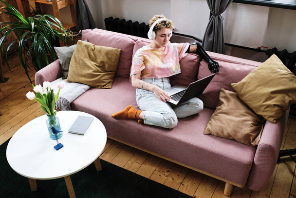 Vista de ángulo alto de la mujer joven con el brazo protésico en los auriculares inalámbricos sentados en el sofá y ver vídeo en línea en el ordenador portátil en casa - Foto, imagen