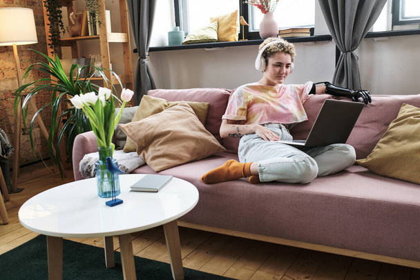 Menina em fones de ouvido com braço protético assistindo vídeo online no laptop enquanto descansa no sofá na sala de estar - Foto, Imagem