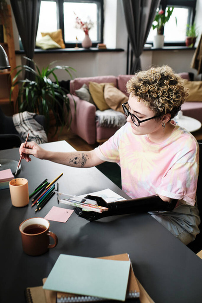 Молода жінка з протезною рукою концентрується на своєму хобі, вона малює картину за столом
 - Фото, зображення