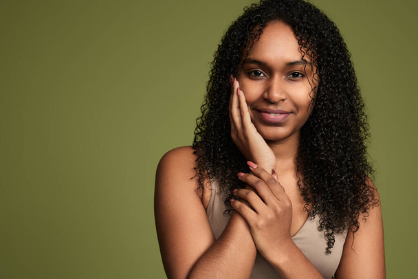 A young woman with long, curly hair is happily smiling and touching clean face with copy space against a green background. - Photo, image