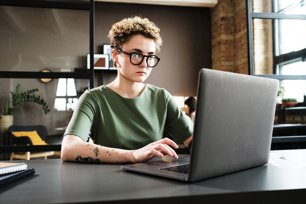 Mujer joven en gafas graduadas concentrándose en su trabajo en línea en el ordenador portátil en su lugar de trabajo en la oficina - Foto, Imagen