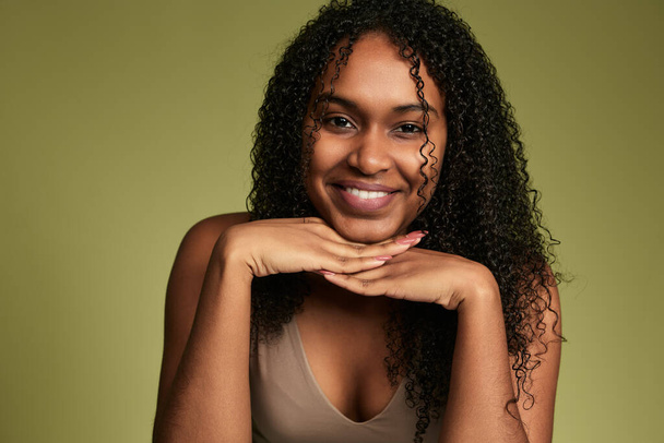 Positive African American female with curly hair and bare shoulders looking at camera while standing on yellow background in studio - Zdjęcie, obraz