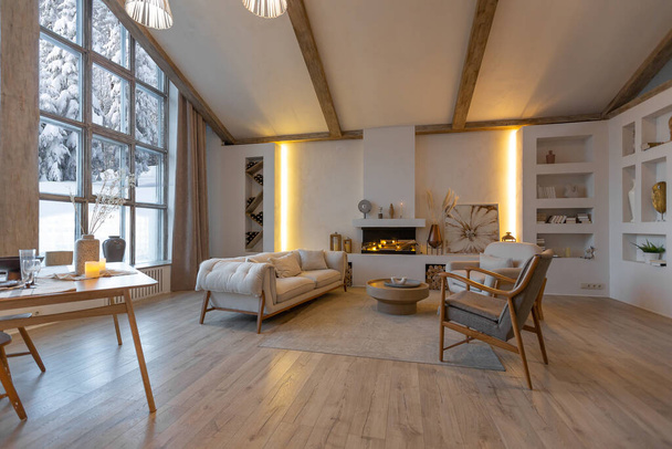 cozy warm home interior of a chic country chalet with a huge panoramic window overlooking the winter forest. open plan, wood decoration, warm colors and a family hearth - Foto, Imagen