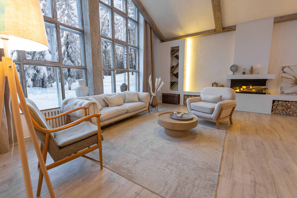 cozy warm home interior of a chic country chalet with a huge panoramic window overlooking the winter forest. open plan, wood decoration, warm colors and a family hearth - Foto, afbeelding