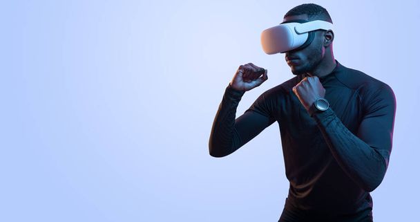 Strong young fit African American male boxer in activewear and VR goggles punching, while experiencing virtual reality against blue background - Fotó, kép