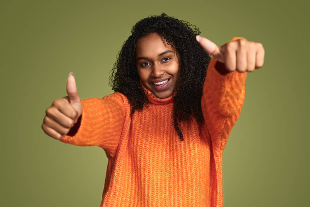 Young carefree ethnic woman wearing vivid orange sweater showing thumbs up and smiling on green background - Fotó, kép