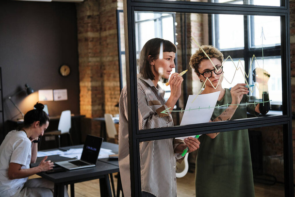 Deux collègues dessinent des graphiques d'entreprise ensemble sur un mur de verre et en discutent pendant la journée de travail au bureau - Photo, image