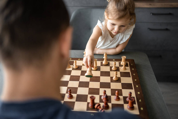 Little girl playing chess with her father at the table in home kitchen. The concept early childhood development and education. Family leisure, communication and recreation. High quality photo - Photo, image