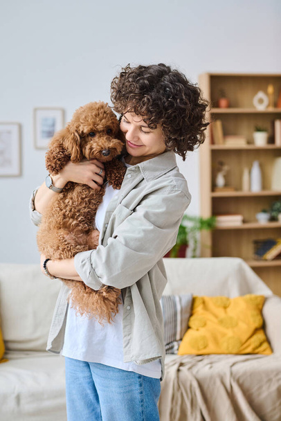Κάθετη εικόνα της νεαρής γυναίκας που κρατά το σκυλί στην αγκαλιά της και το αγκαλιάζει ενώ στέκεται στο δωμάτιο - Φωτογραφία, εικόνα