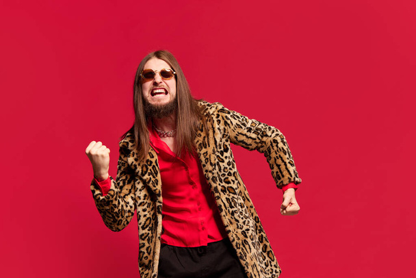 Portrait of young man with long hair posing in stylish fur coat and sunglasses against red studio background. Emotionally shouting. Concept of emotions, facial expression, lifestyle - Photo, Image