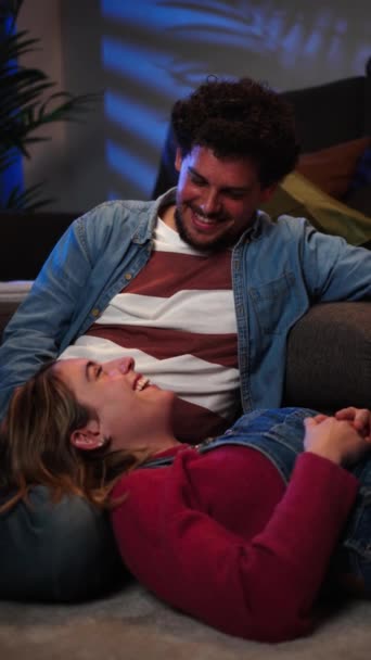 Vertical view of young caucasian couple smiling and looking at each other, resting sitting on the carpet on the floor at home and leaning on the sofa on a romantic date Husband and wife relaxing. High - Footage, Video