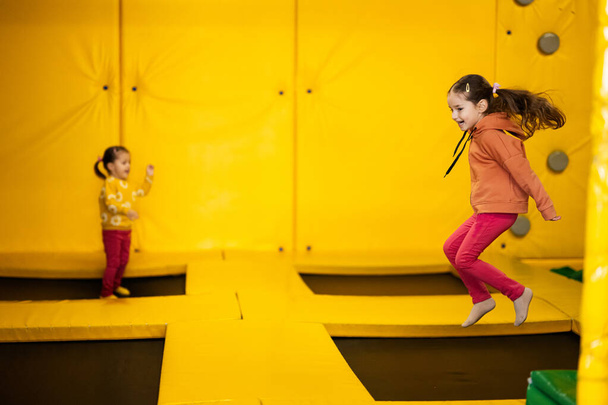 Siblings jumping on trampoline at yellow playground park. Sisters in motion during active entertaiments. - Fotó, kép