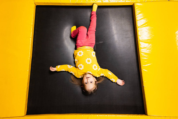 Baby girl kid lies upside down on trampoline at playground park. Child in motion during active entertaiments. - Photo, Image