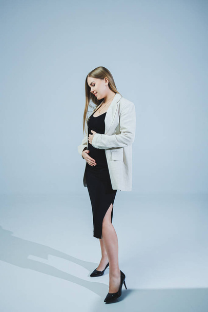 A young woman in a black top, skirt and beige jacket stands on a white background and hugs her pregnant belly. Attractive pregnant woman on a white background. Casual clothes for stylish and fit women - Photo, Image