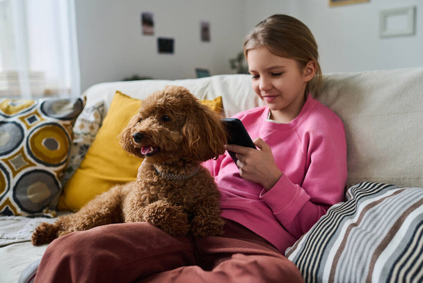Bambina seduta sul divano con cane e giocare nel suo smartphone durante il tempo libero a casa - Foto, immagini