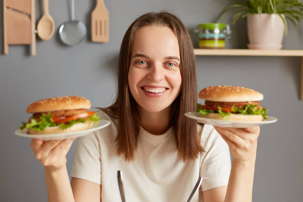 Indoor shot of smiling cheerful Caucasian woman holding two delicious fresh burgers, posing in kitchen, breaking diet, feels extremely hungry, looking at camera with happy face. - Foto, Bild