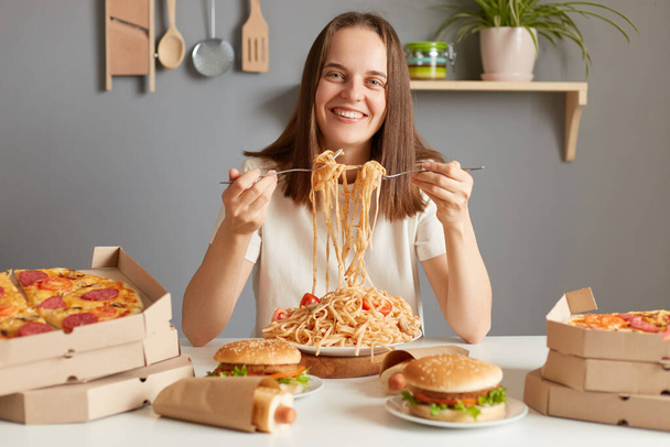 Portrait of smiling happy positive woman wearing white T-shirt sitting at table in kitchen, enjoying tasty dinner, having cheat meal, breaking diet. - Valokuva, kuva