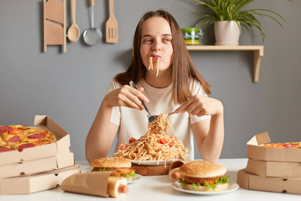Zdjęcie zadowolonej zachwyconej kobiety noszącej biały T-shirt siedzącej przy stole w kuchni i jedzącej makaron, pozującej z ustami pełnymi makaronu, łamiącej dietę, jedzącej z satysfakcją. - Zdjęcie, obraz
