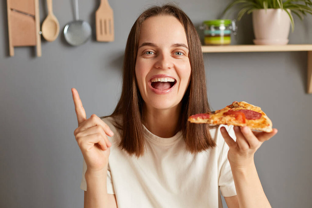 Portrait of amazed happy woman holding piece of pizza, feels excited wearing casual white T-shirt posing in kitchen, raised finer up, eating pizza, having idea how to burn calories. - Foto, immagini