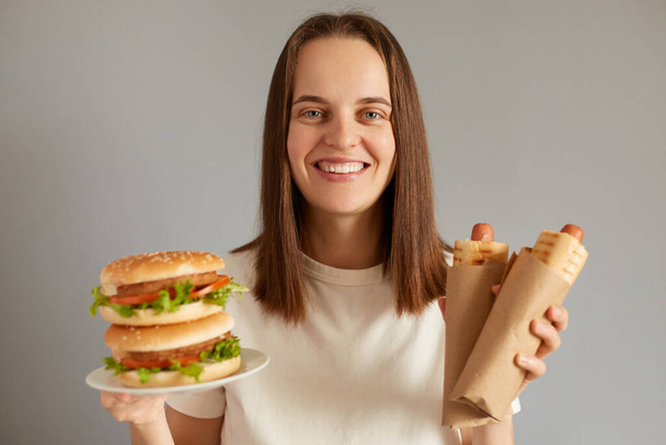 Indoor shot of smiling attractive brown haired woman holding big delicious fresh burger and two hot dogs, enjoying fast food, looking at camera and smiling. - Foto, Imagem