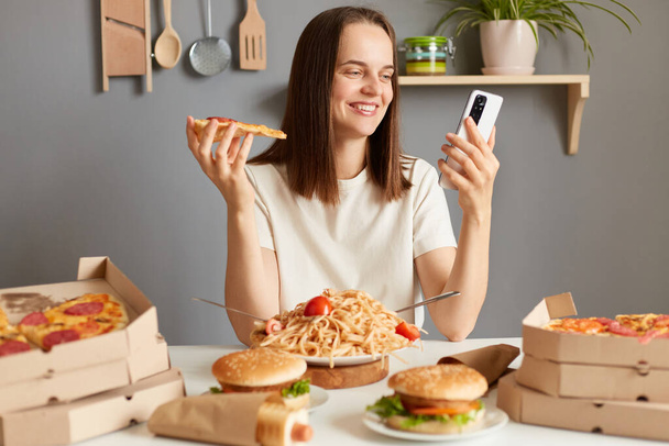 Portrait of attractive charming woman with brown hair sitting in kitchen and using mobile phone while eating tasty pizza, chatting with friends, left positive feedback delivery service. - Photo, Image