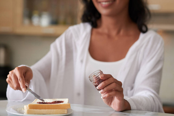 Woman speading homemade jelly from small jar on toast when making breakfast in the morning - Photo, image